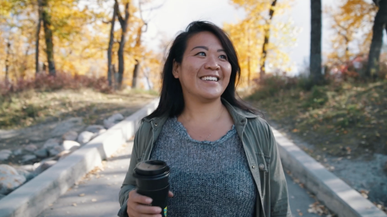 Kate Mak is standing on a walking path with trees along the pathways. She is smiling, holding a to-go coffee cup.