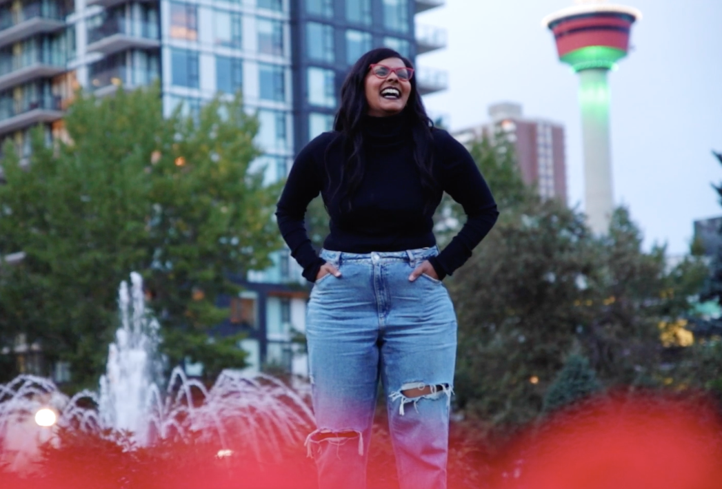 Subiksha Subramani mid-laugh, wearing a black long-sleeve top, light denim jeans, and red glasses, with the green-lit Calgary Tower in the background at Central Memorial Park.